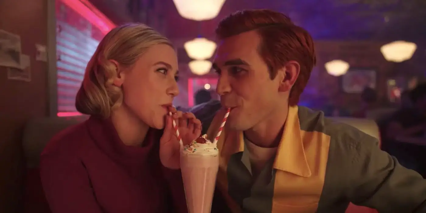 Betty (Lili Reinhart) y Archie (KJ Apa) sonriendo y bebiendo del mismo batido con dos pajitas en un restaurante en la temporada 7 de Riverdale