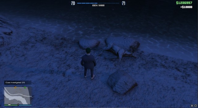 Un personaje de GTA Online, con pelo verde y traje azul, de pie junto a un ciervo muerto.  El ciervo está al lado del río, contra unas rocas. 