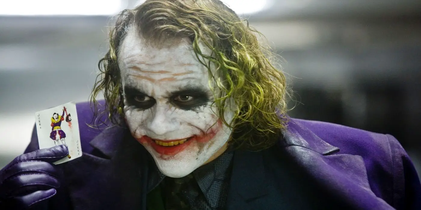 Heath Ledger como The Joker sonriendo mientras muestra una carta comodín en The Dark Knight